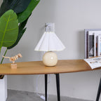 In this photo, a Clérachic table lamp with a white pleated shade stands on an oval wooden table, plugged into a wall socket above it.