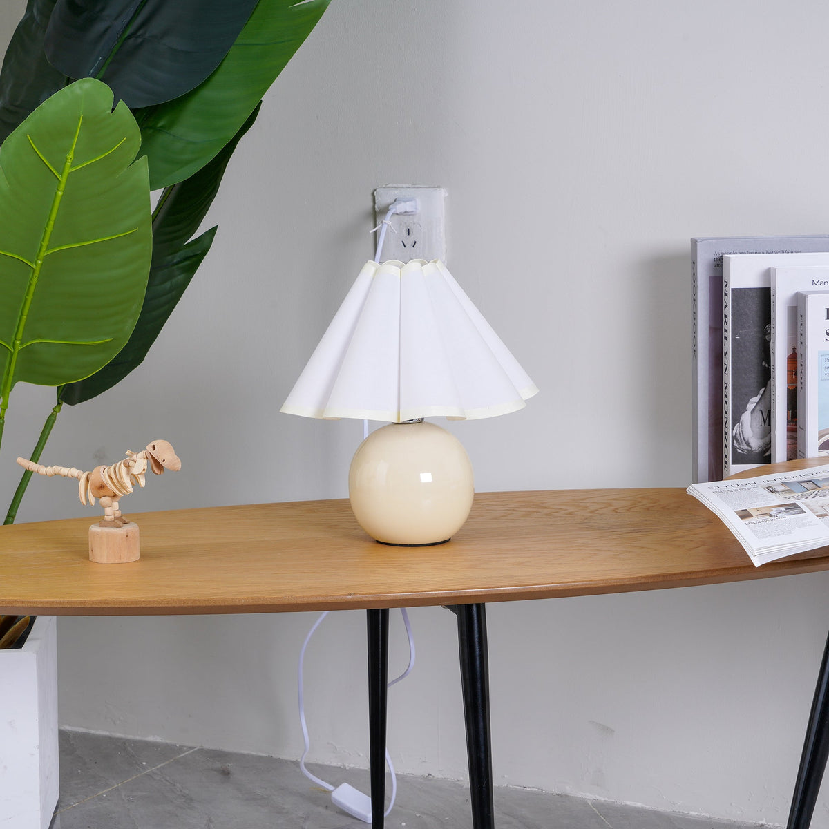 In this photo, a Clérachic table lamp with a white pleated shade stands on an oval wooden table, plugged into a wall socket above it.