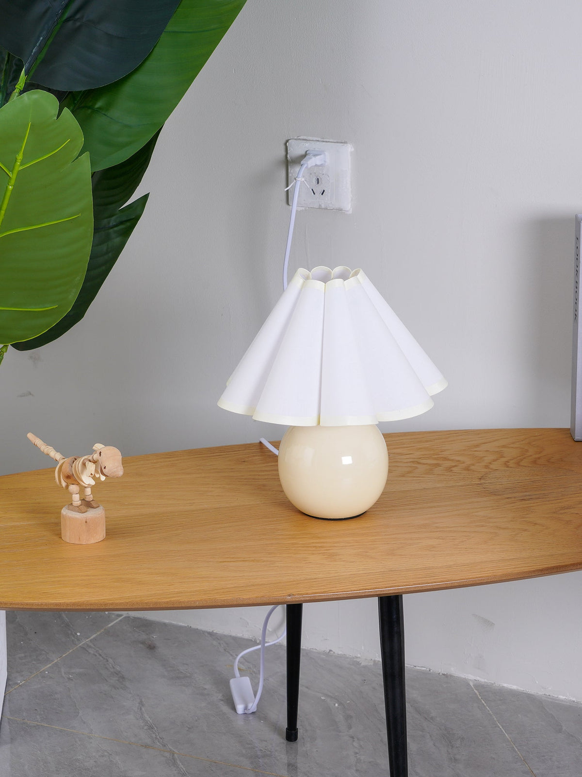 This Clérachic table lamp with a white pleated shade stands on an oval wooden table, with a toy dinosaur and a large leaf at its left.