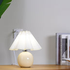 Close to the camera, this Clérachic table lamp with a white pleated shade glows beside a row of books, plugged into the wall.