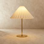 Clérachic table lamp with a cream pleated conical shade, glowing warm on a slim brass stem and round brass base against limewash.