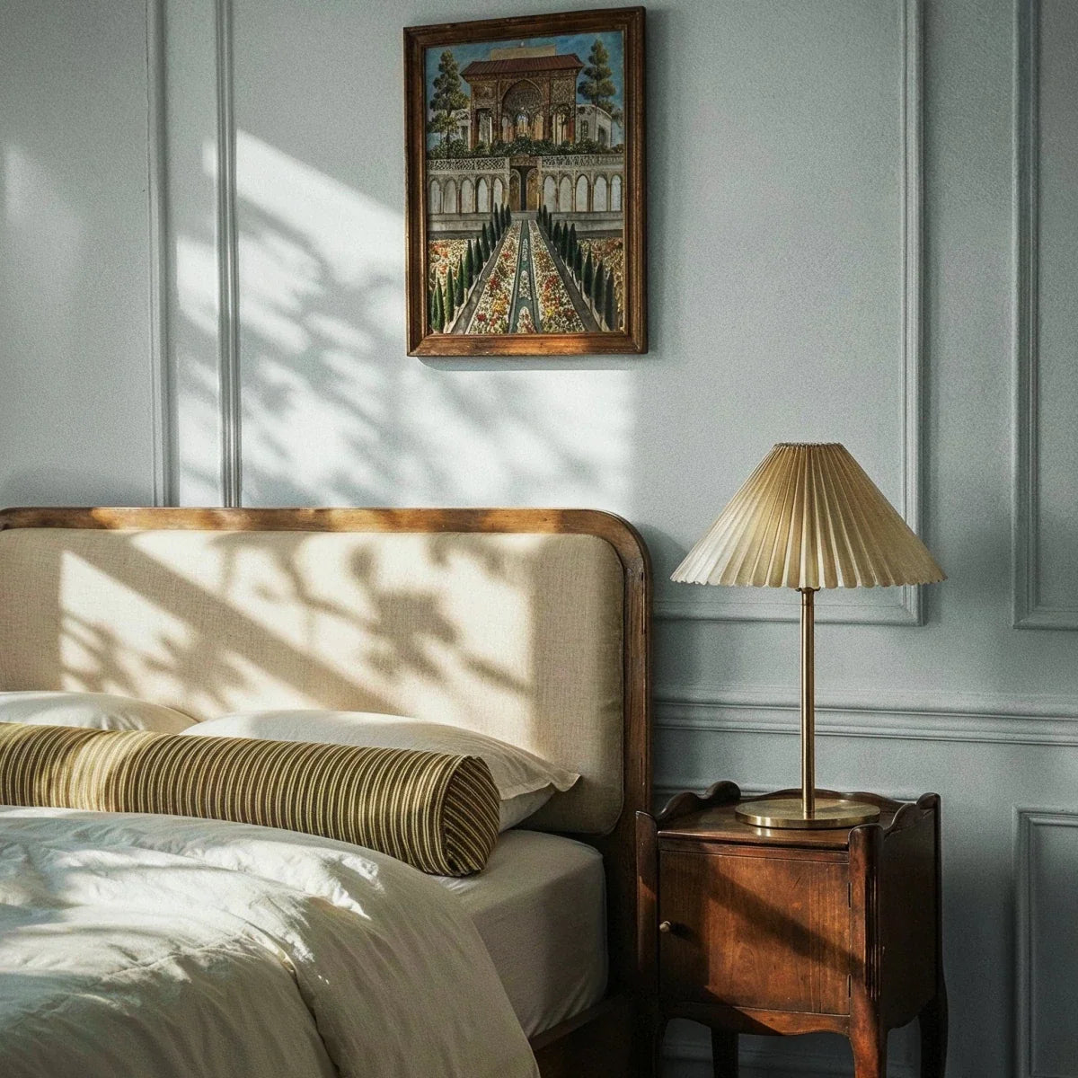 Clérachic pleated-shade table lamp on a slim brass stem, set on a dark wood bedside cabinet beside a linen headboard and panelled wall.