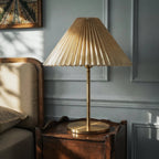 Clérachic pleated linen-toned shade on a slim brass stem and round base, on a scalloped dark wood bedside table in morning light.