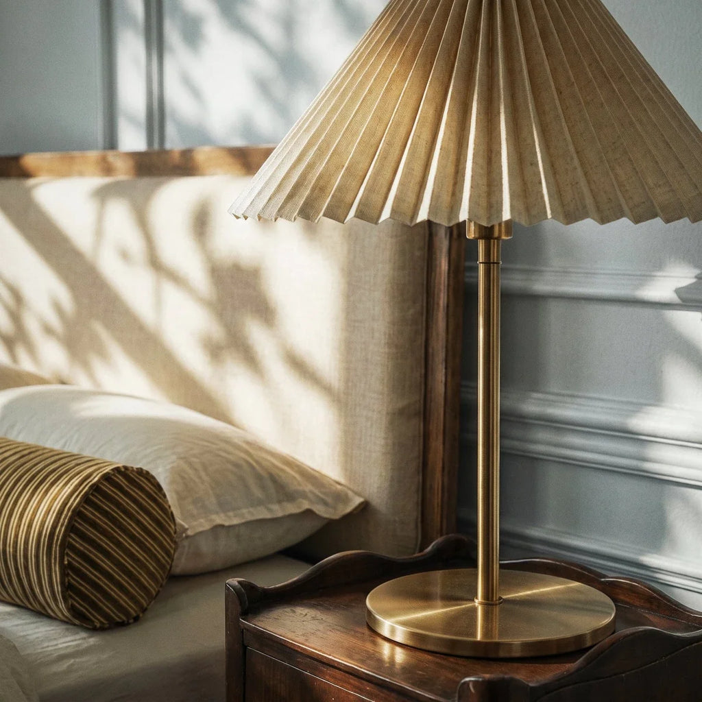 Close view of a Clérachic pleated shade and slim brass stem on a round base, standing on a wooden bedside table beside a striped bolster.