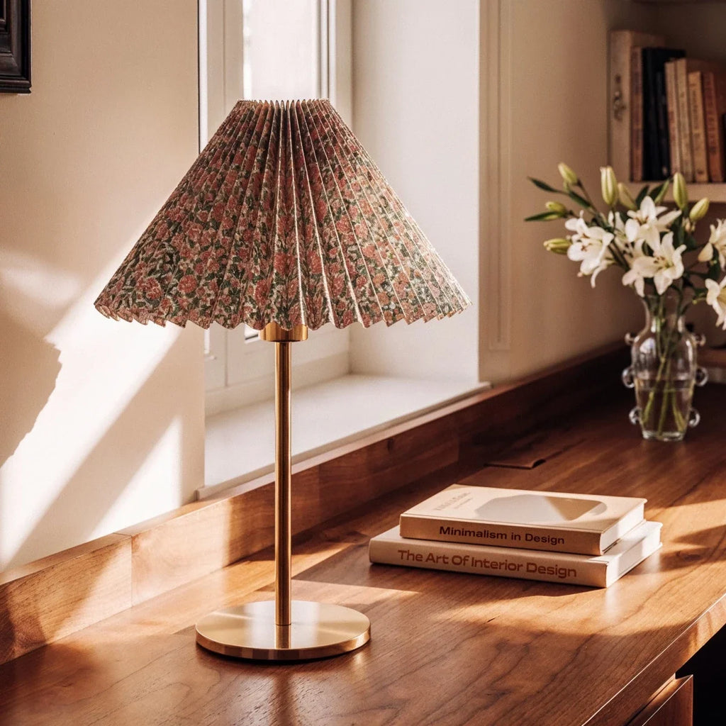 Clérachic floral pleated-shade table lamp with brass stem and round base on a walnut desk, beside stacked books and white lilies.