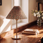 Clérachic floral pleated-shade table lamp with brass stem and round base on a walnut desk, beside stacked books and white lilies.