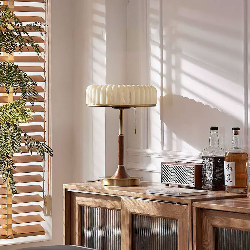 This Clérachic table lamp stands on a wooden cabinet by a window, sunlight through blinds falling across the pleated beige shade.