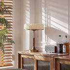 This Clérachic table lamp stands on a wooden cabinet by a window, sunlight through blinds falling across the pleated beige shade.