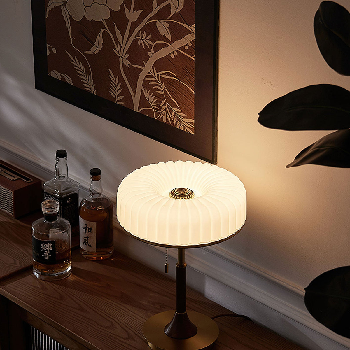 Glowing warm white, this Clérachic table lamp sits on a wooden console beneath a botanical painting, with bottles at its side.