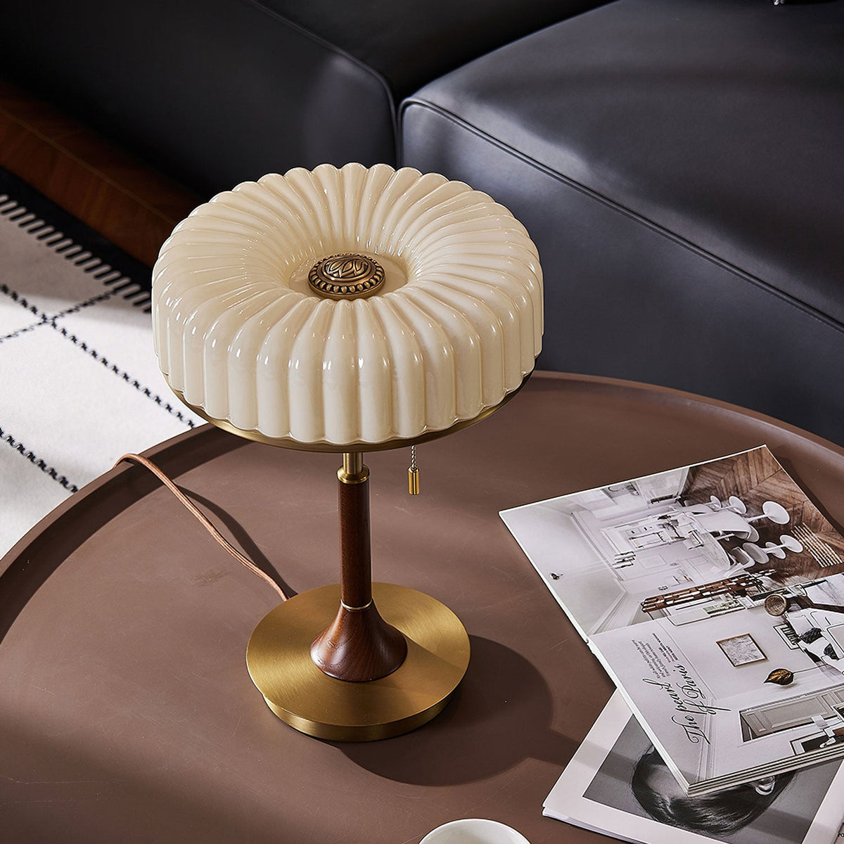 Top-down view of this Clérachic table lamp on a dark round table, the pleated shade above gold base and walnut stem, with magazines nearby.