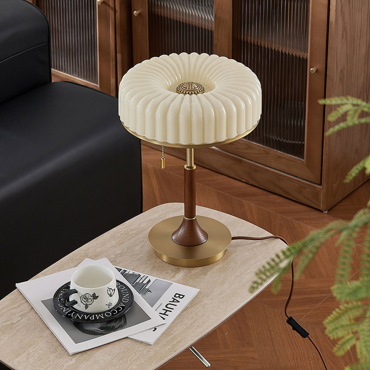 In this photo, a Clérachic table lamp on a pale stone side table beside a black sofa, with its cord trailing off the edge and a fern leaf in frame.