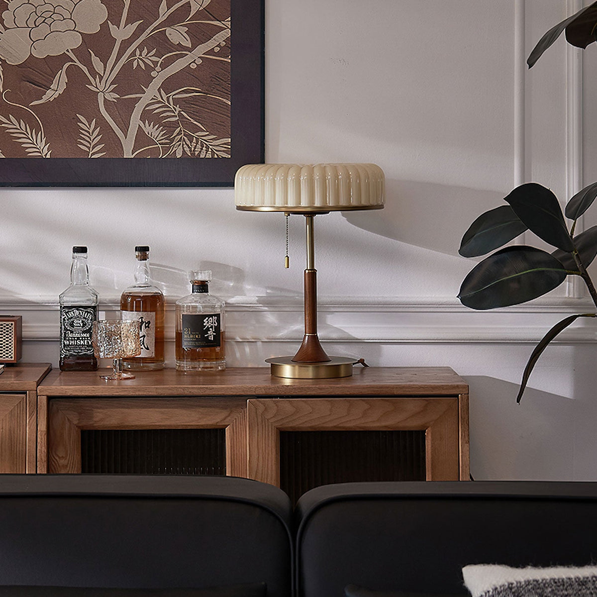 Unlit Clérachic table lamp on a walnut sideboard, the pleated beige shade above a short wooden stem and gold base, with a plant to the right.