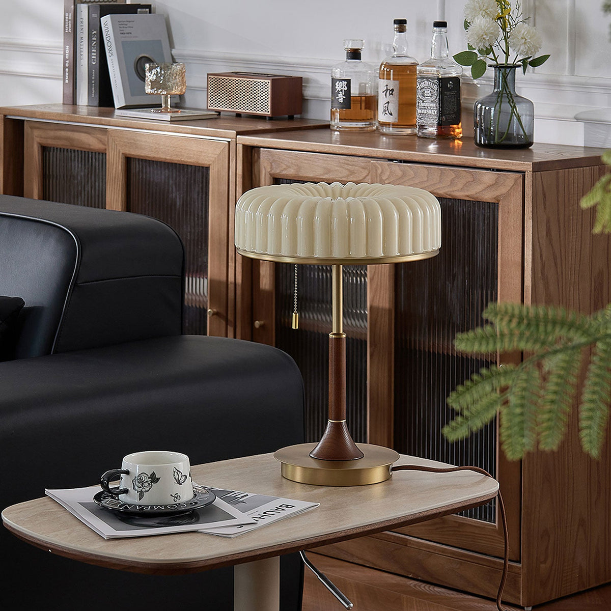 Pictured here, a Clérachic table lamp on a wooden cabinet behind a stone side table, with a cup on a magazine and a black sofa arm at the left.