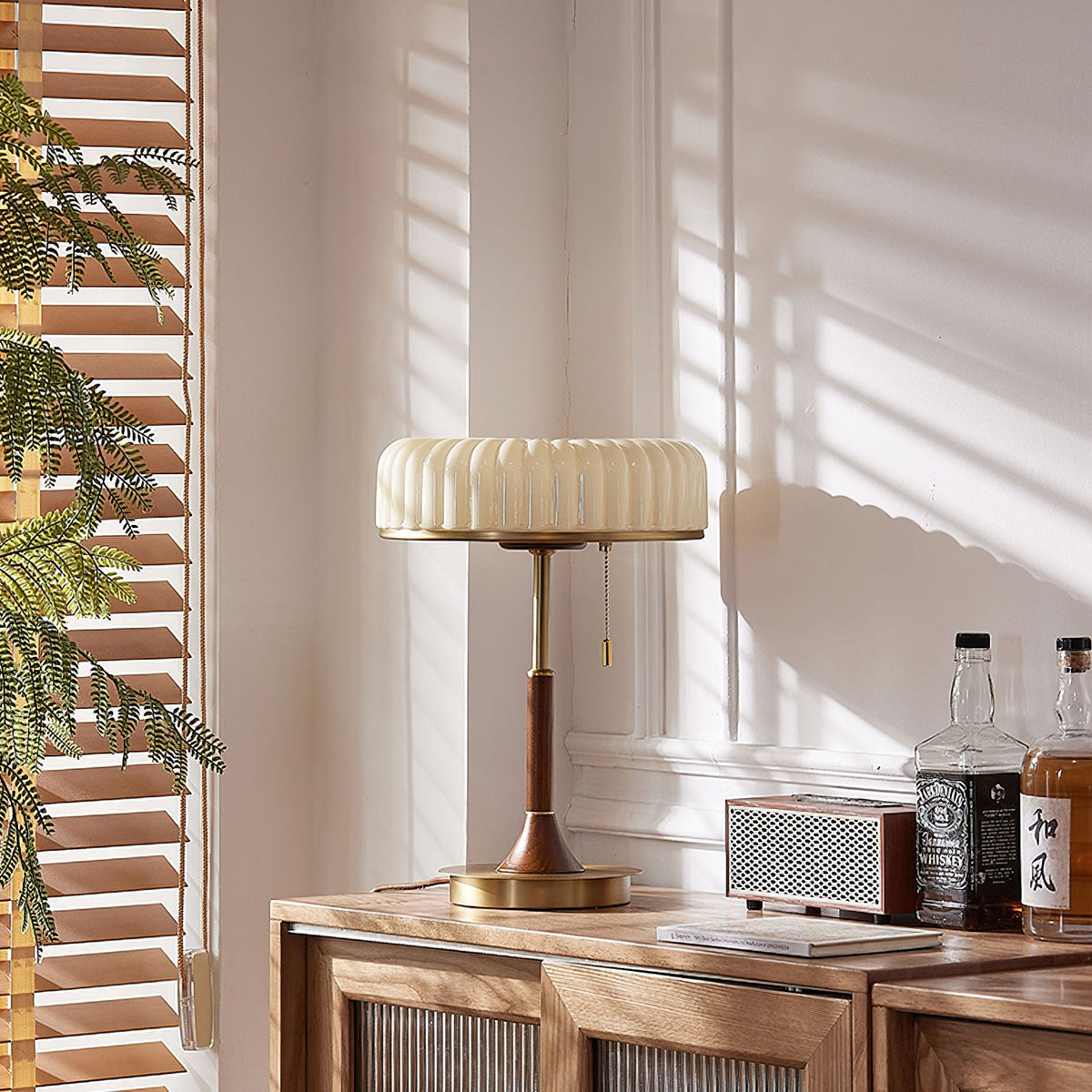 Sunlit corner with a Clérachic table lamp on a walnut cabinet, shadows of window blinds across the wall and a small radio beside it.