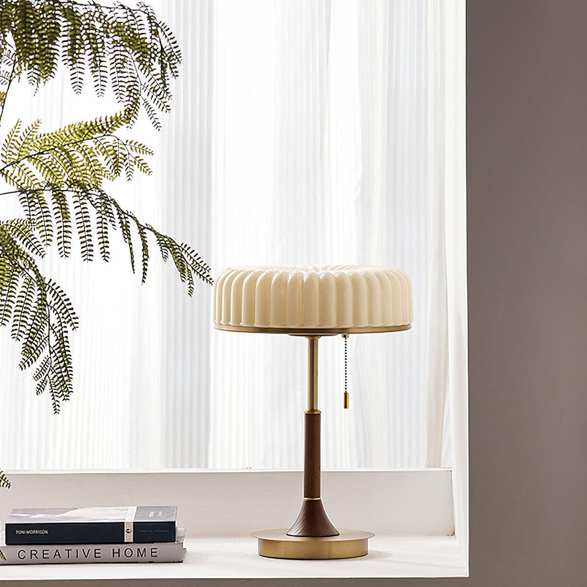 A Clérachic table lamp on a white windowsill with sheer curtains behind it, a feathery plant to the left and two books at its side.