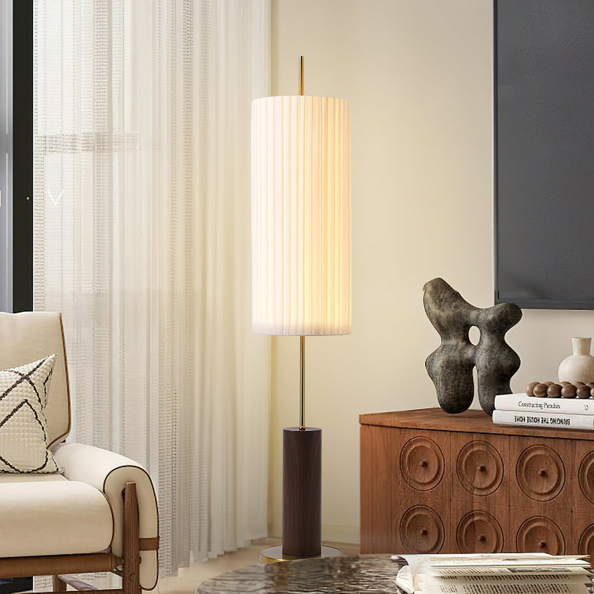 Lit pleated shade of this Clérachic floor lamp beside a carved walnut cabinet and a cream armchair, with sheer curtains behind.