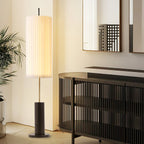 A Clérachic floor lamp glowing in a corner beside a slatted black sideboard, a tall plant and a mirror on the wall.
