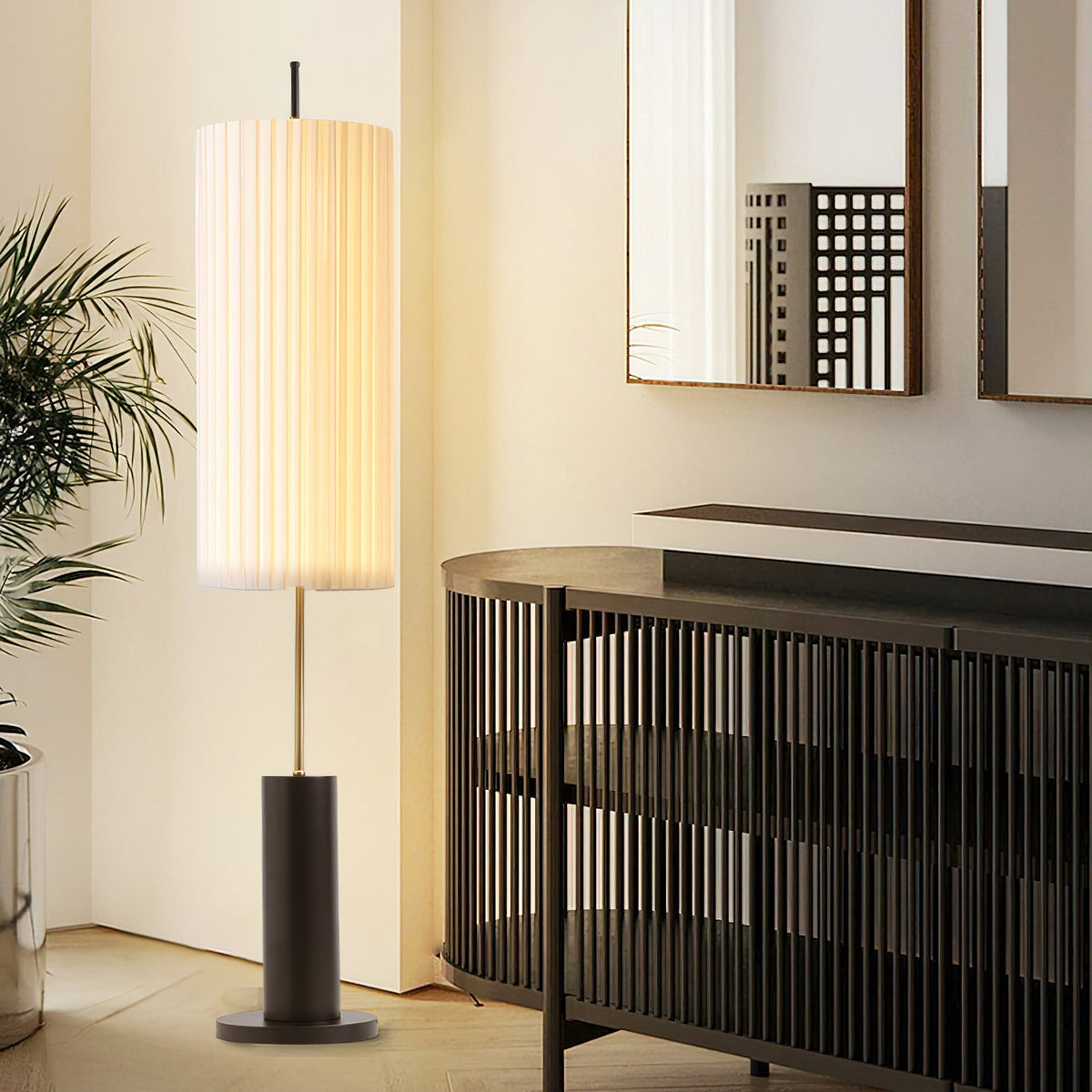 A Clérachic floor lamp glowing in a corner beside a slatted black sideboard, a tall plant and a mirror on the wall.