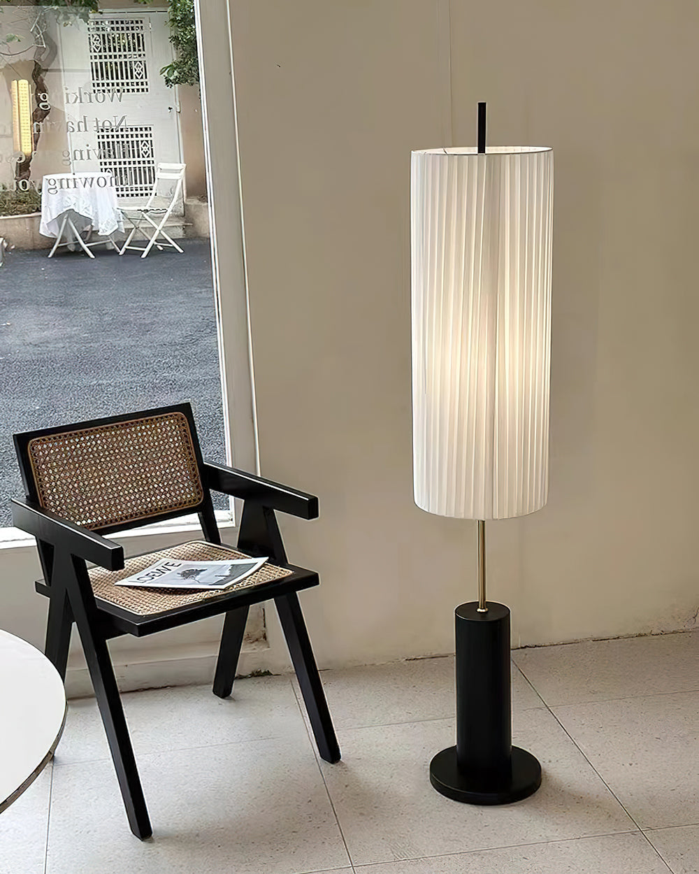 A Clérachic floor lamp with a black base beside a cane-seated black armchair, in front of a glass door on a tiled floor.