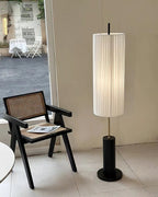 A Clérachic floor lamp with a black base beside a cane-seated black armchair, in front of a glass door on a tiled floor.