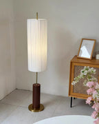 Pleated Clérachic floor lamp lit in a tiled corner beside a cane-fronted cabinet, with a brass foot under its walnut-colour base.