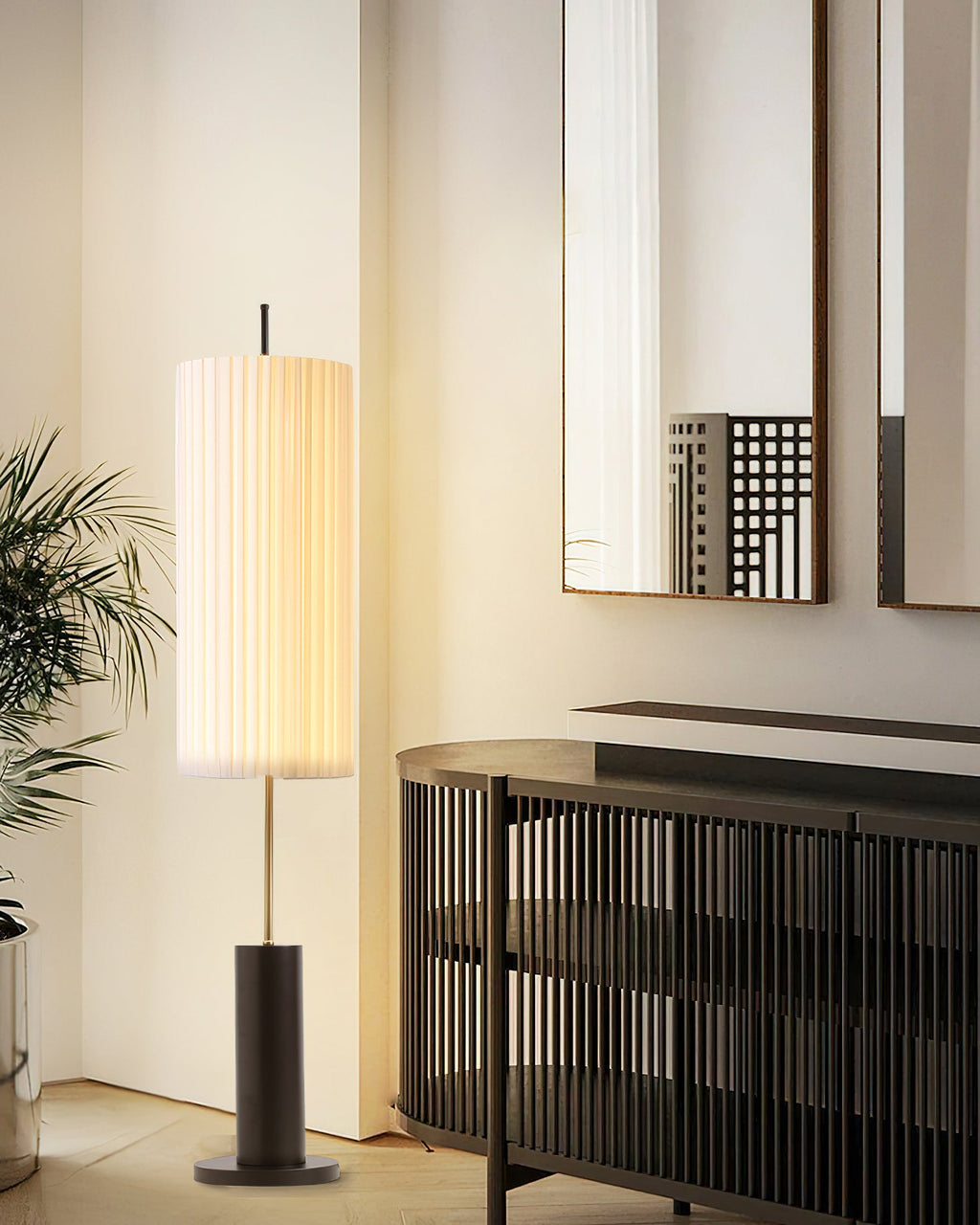 Lit Clérachic floor lamp beside a tall mirror, a palm and a slatted dark sideboard, with a black base under the pleated shade.