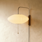 Clérachic wide saucer-shaped pleated fabric wall lamp, lit warm, on a chrome arm with a wooden mount and pull chain.