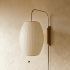 Clérachic barrel-shaped pleated white fabric wall lamp, switched off, on a chrome arm with a wooden mount and cord.