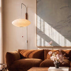 Close view of a Clérachic saucer-shaped pleated wall lamp with a red pull bead, hanging over a velvet sofa.