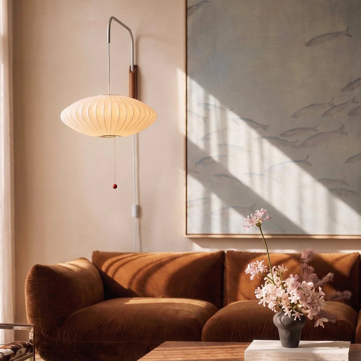 Close view of a Clérachic saucer-shaped pleated wall lamp with a red pull bead, hanging over a velvet sofa.