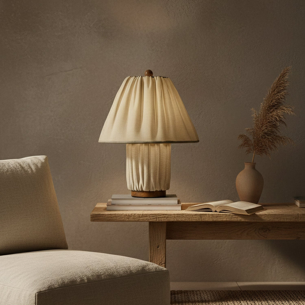 A Clérachic pleated linen lamp glows softly on a wooden console beside a stack of books and dried pampas grass.