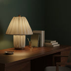 A lit Clérachic pleated table lamp on a dark wood desk, with books and a brass pen pot against a deep green wall.