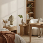 This Clérachic pleated lamp sits on a round bedside table in a calm bedroom, with a plant, a vase and a shelving unit behind.