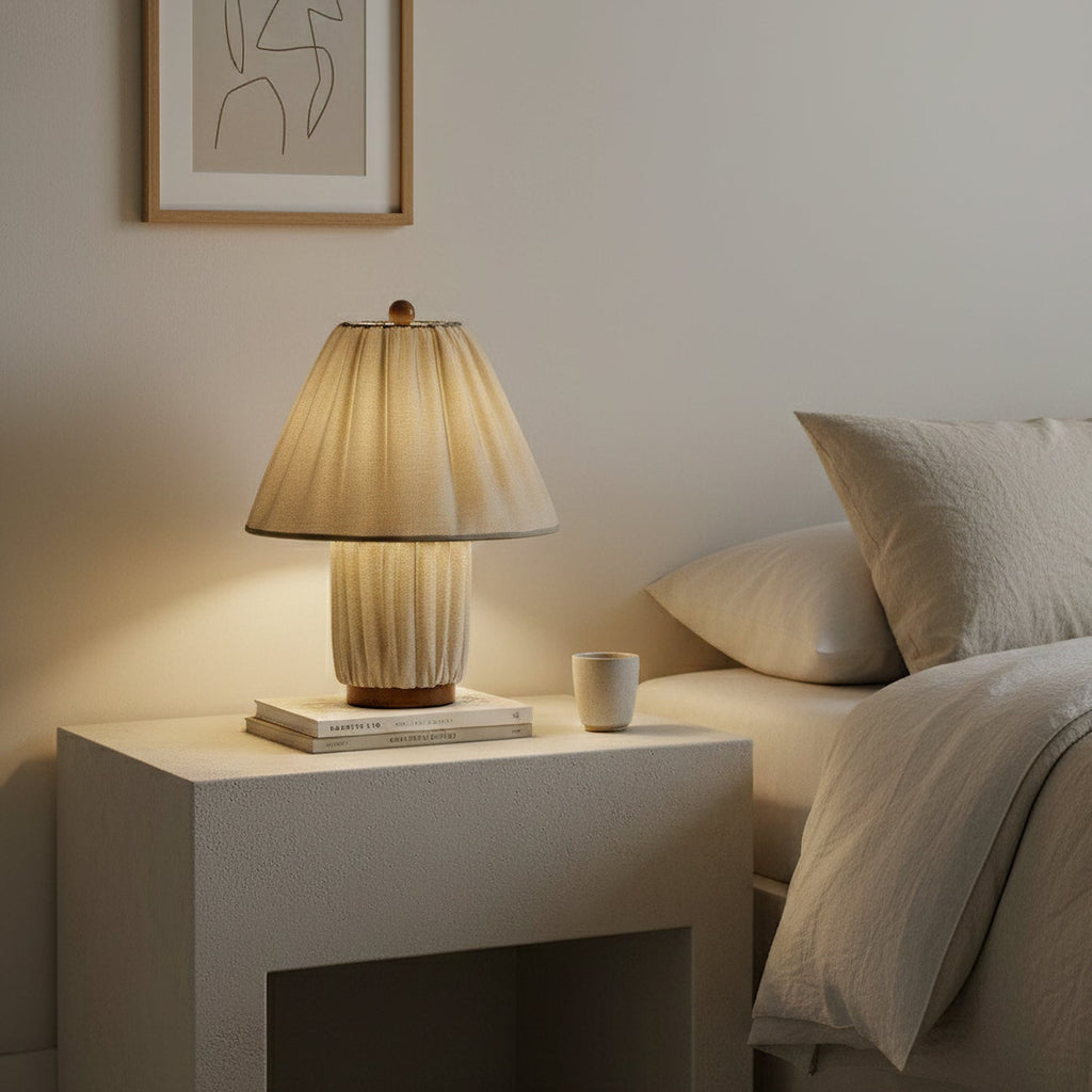 A Clérachic pleated linen lamp lights a white bedside block beside a bed, with a mug and a small stack of books.