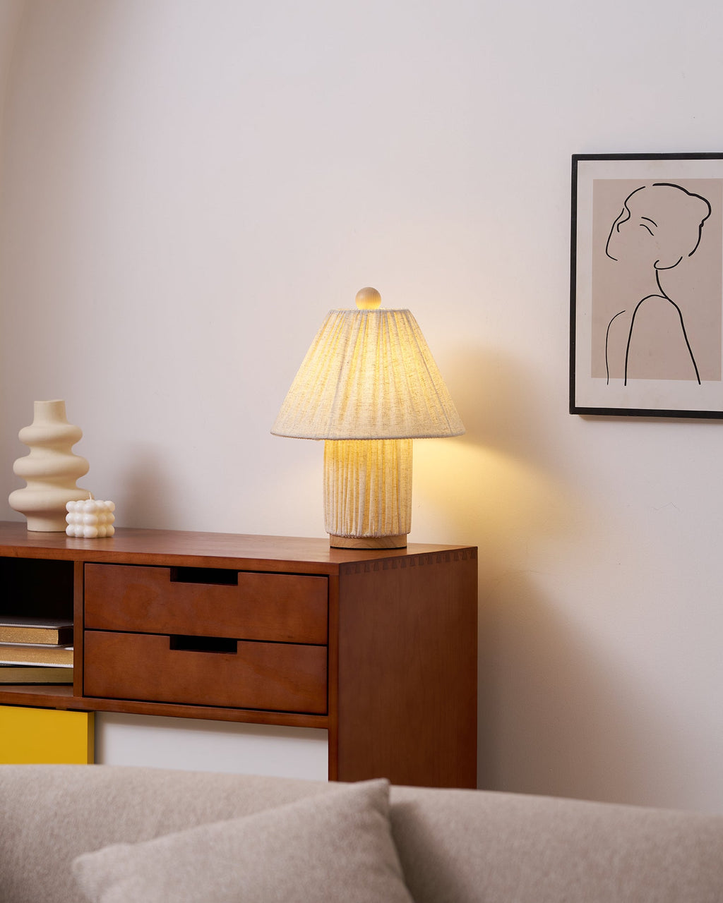 A lit Clérachic pleated lamp on a walnut sideboard with two drawers, beside a cream sculptural vase and a framed line drawing.