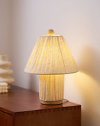 Close view of the lit shade of this Clérachic pleated lamp, with its wooden finial, on the corner of a walnut sideboard.