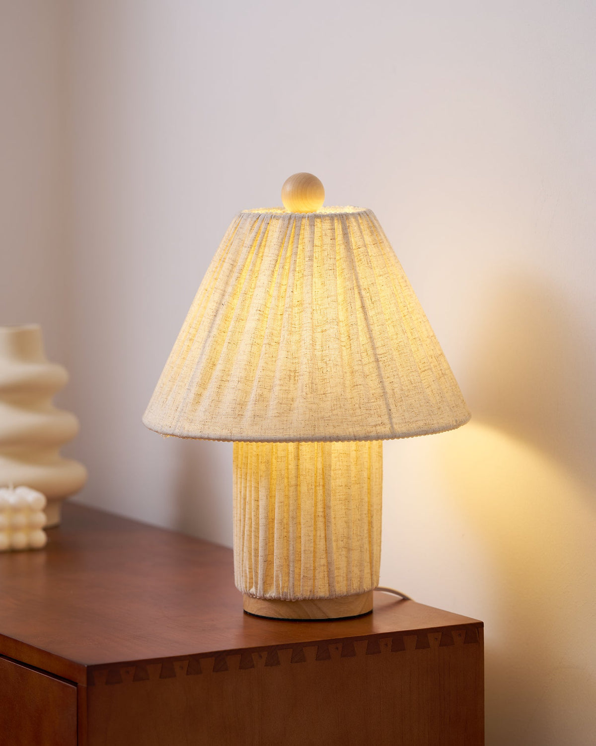 Close view of the lit shade of this Clérachic pleated lamp, with its wooden finial, on the corner of a walnut sideboard.