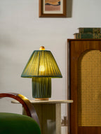 Dark green Clérachic pleated lamp lit on a travertine side table, with a rattan cabinet and a green velvet chair nearby.