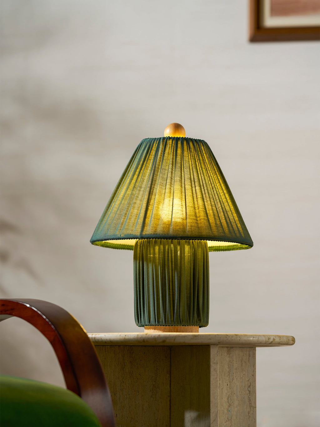 Close view of a lit dark green Clérachic pleated lamp on a travertine side table, beside a rattan-fronted cabinet.