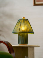 Close view of a lit dark green Clérachic pleated lamp on a travertine side table, beside a rattan-fronted cabinet.