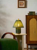 A smaller view of the dark green Clérachic pleated lamp, lit, on a stone table by a rattan cabinet and a framed print.