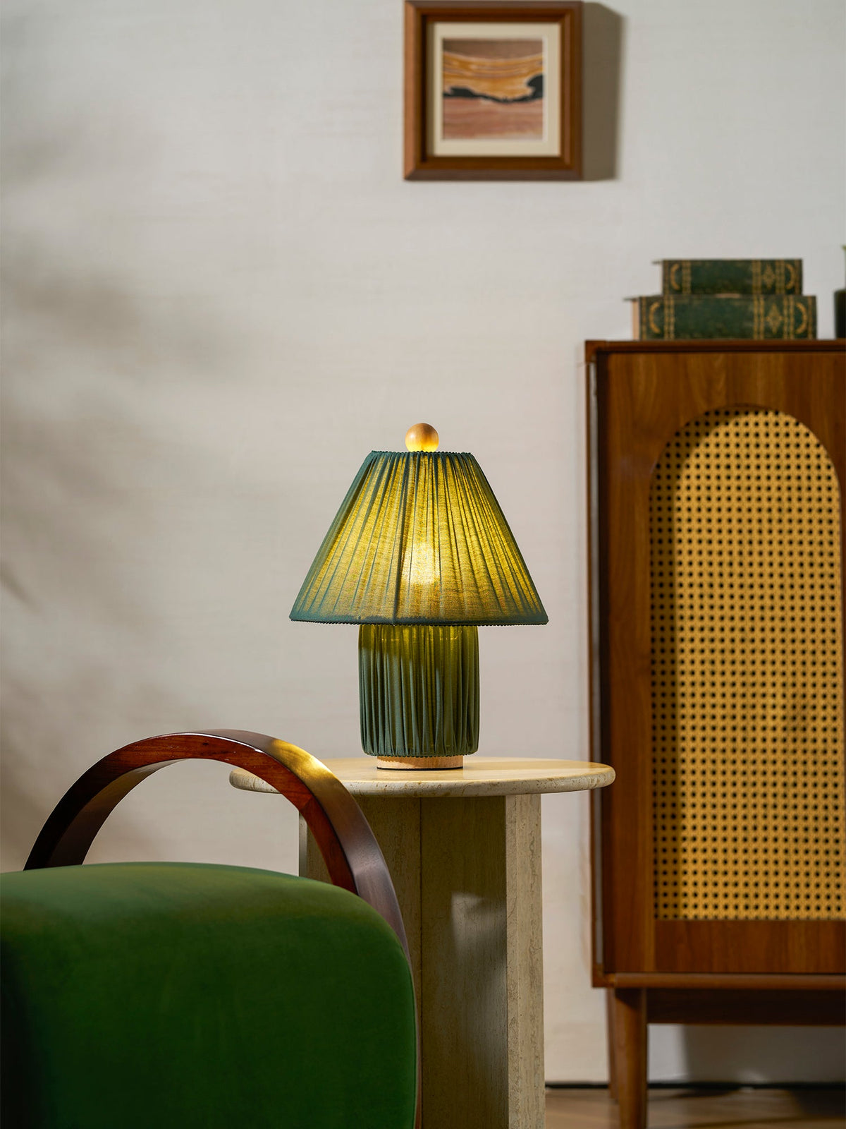 A smaller view of the dark green Clérachic pleated lamp, lit, on a stone table by a rattan cabinet and a framed print.