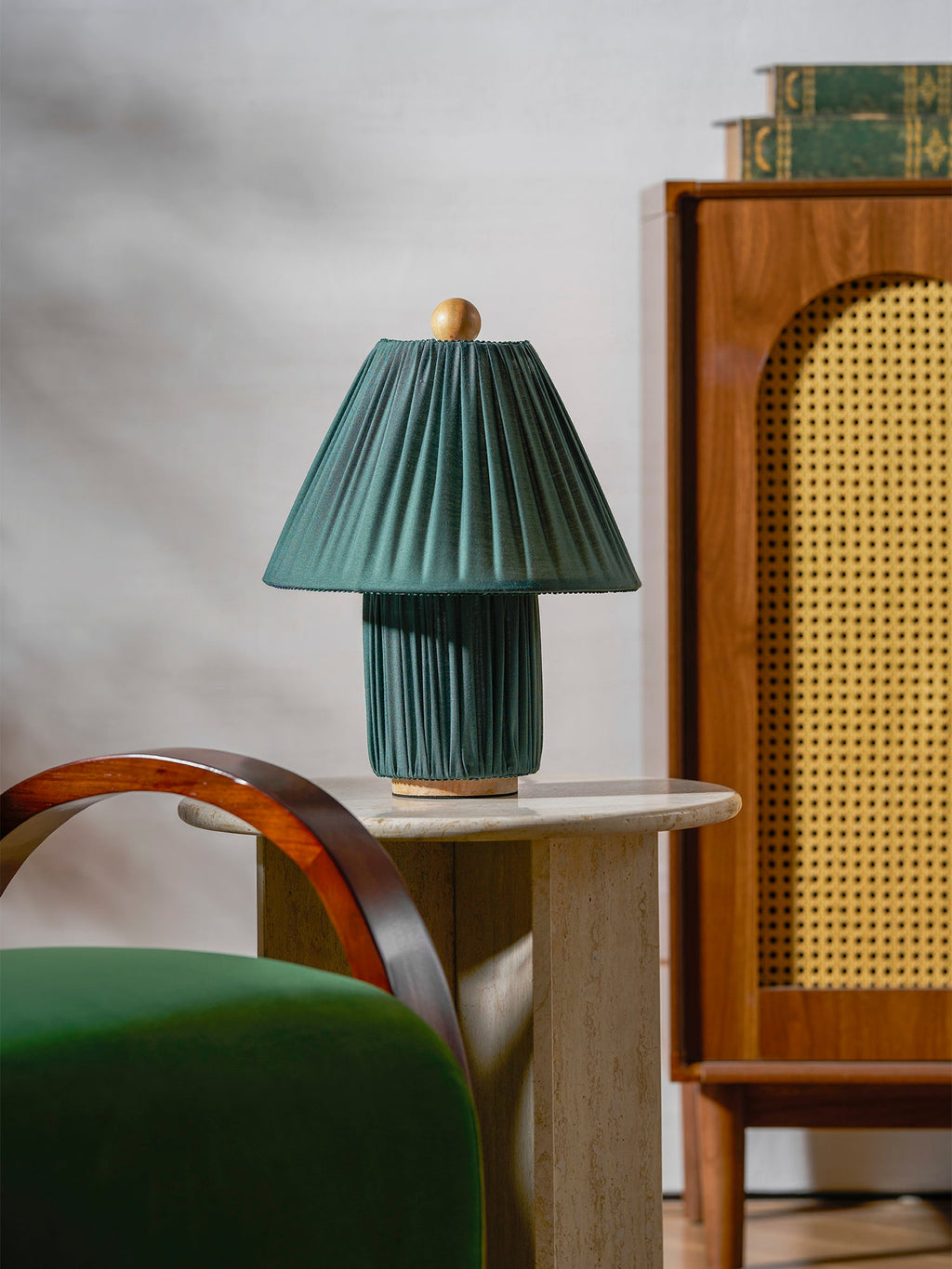 A teal-green version of this Clérachic pleated lamp, unlit, on a round stone table beside a rattan cabinet and green chair.