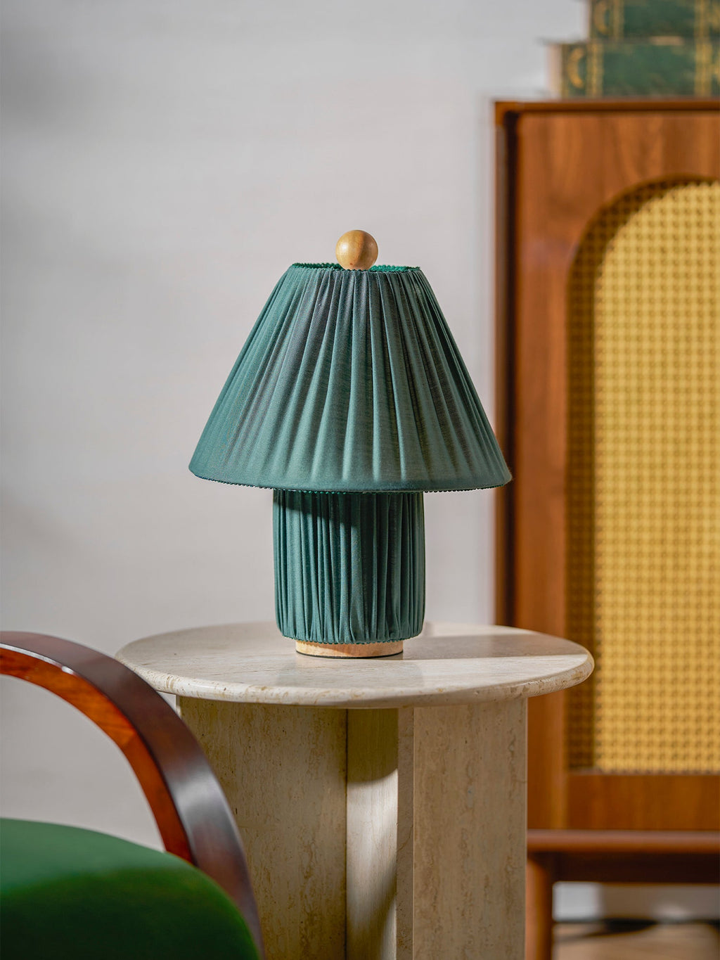 Close view of an unlit teal-green pleated lamp from Clérachic on a round stone table, with a rattan cabinet behind.