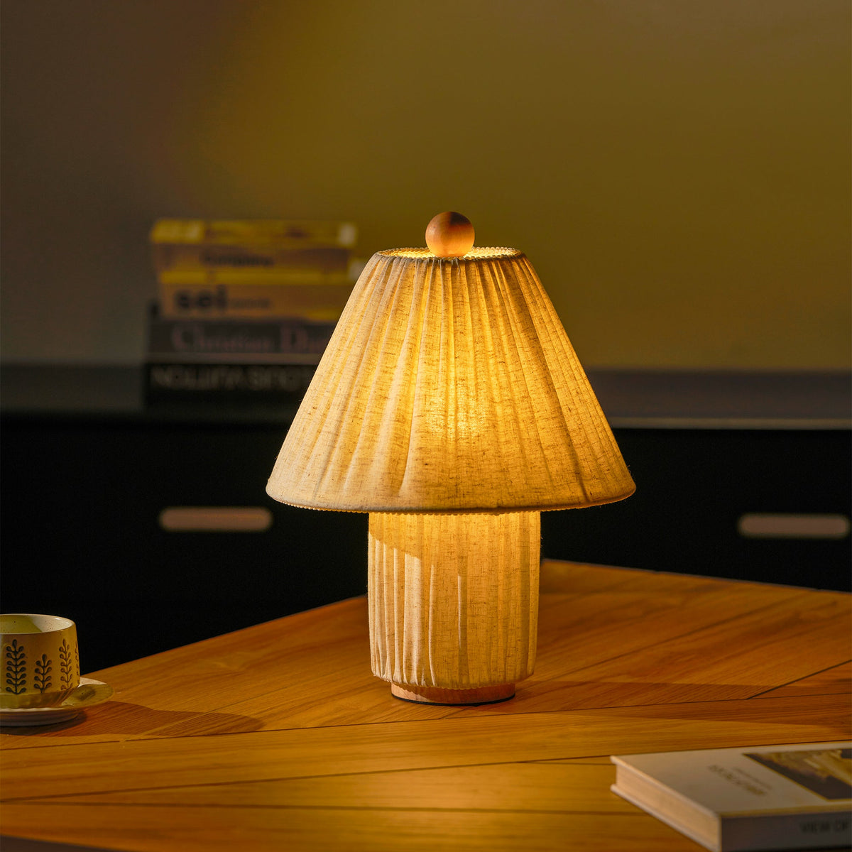 A Clérachic pleated linen lamp glows amber on a wooden table, with a cup and saucer, books and a dark cabinet behind.