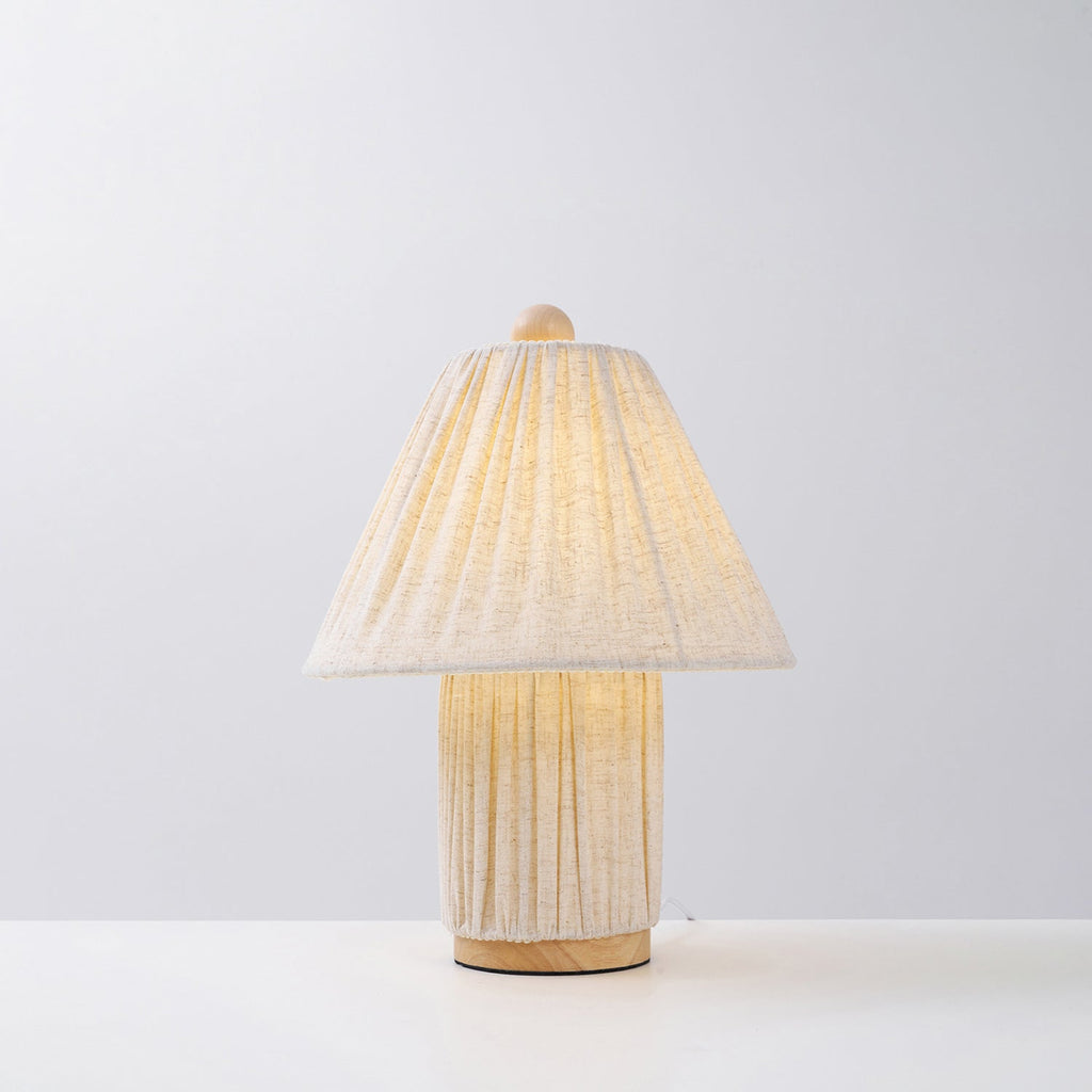 Lit Clérachic linen lamp with a pleated shade and a matching pleated base on a wooden foot, against a plain pale backdrop.