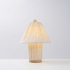 Lit Clérachic linen lamp with a pleated shade and a matching pleated base on a wooden foot, against a plain pale backdrop.