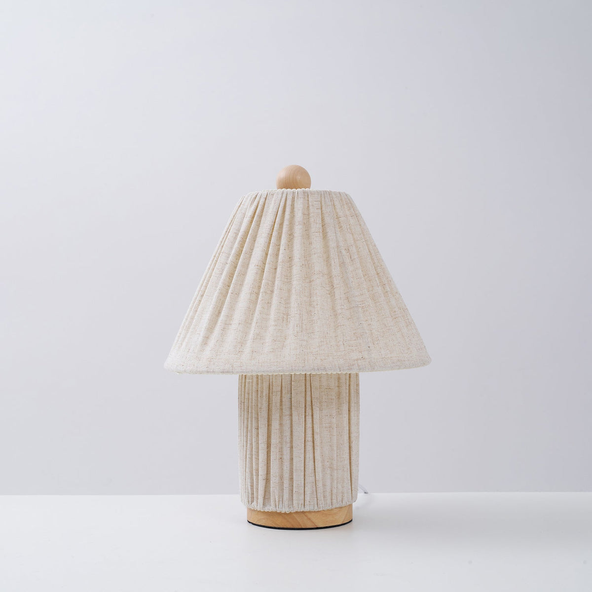 Unlit Clérachic pleated linen lamp with a wooden foot, standing on a white surface against a pale grey wall.