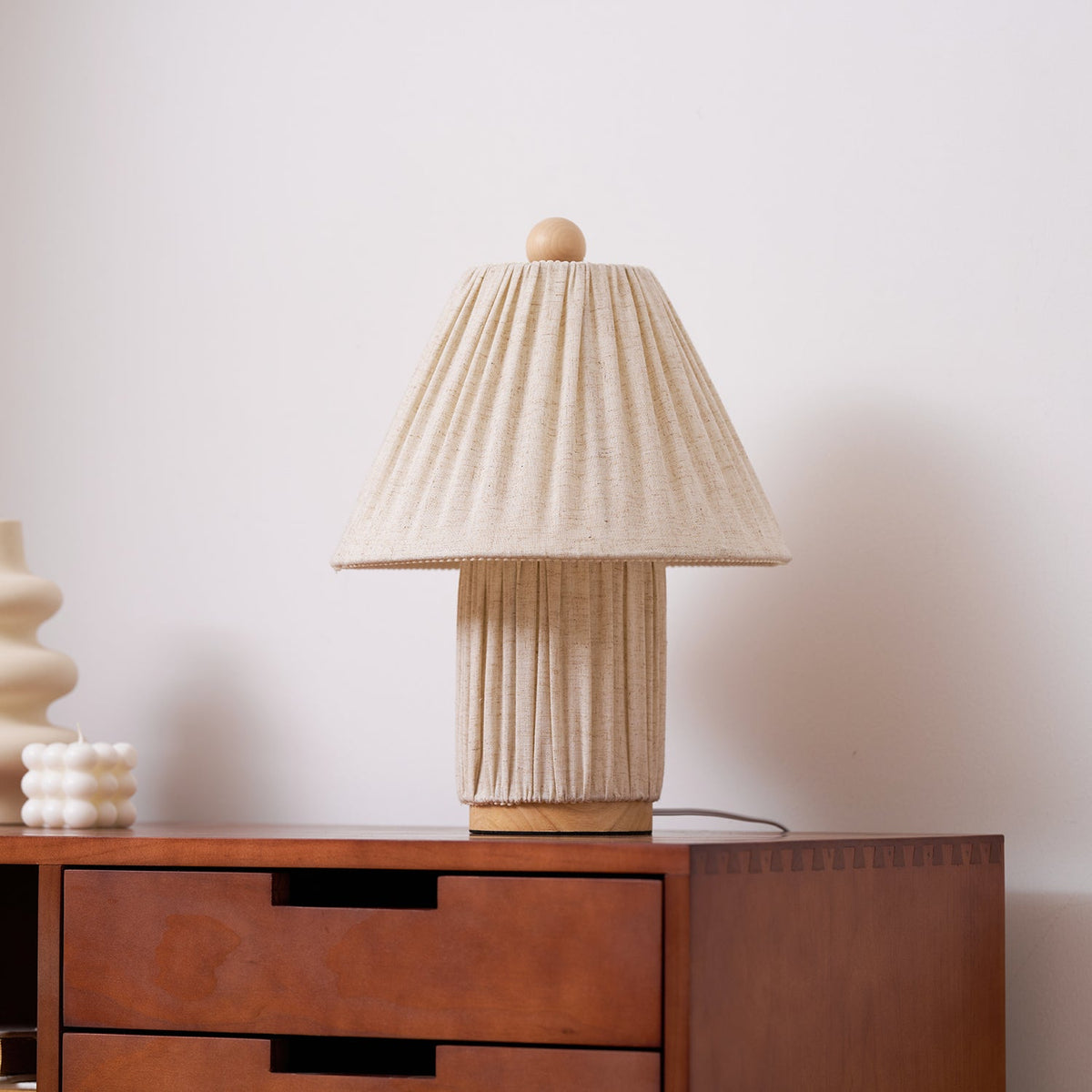 Pleated linen shade and base of this Clérachic lamp, unlit, on a drawered walnut sideboard with a cream vase to the left.