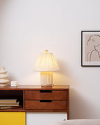 A glowing Clérachic pleated lamp on a walnut sideboard with a yellow panel, beside a vase and a line-drawing frame.